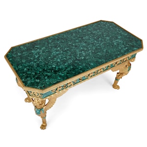 Puede incluir: Mesa de centro rectangular con tapa verde malaquita y ribetes dorados. La mesa presenta patas doradas ornamentadas con figuras aladas y patas de garra. La mesa mide aproximadamente 152 cm de largo.