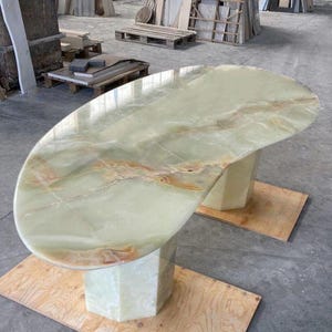 May include: An oval dining table crafted from light green onyx stone with natural tan and brown veining throughout the surface. The table features a smooth, polished top supported by two matching hexagonal pedestals made of the same green onyx.