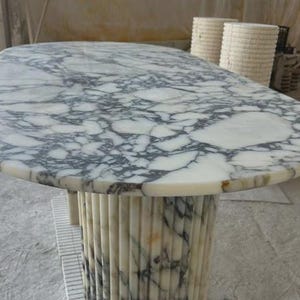 Puede incluir: Mesa ovalada de mármol con tapa veteada en blanco y gris. La base es una columna estriada de mármol a juego. La mesa es un mueble único.