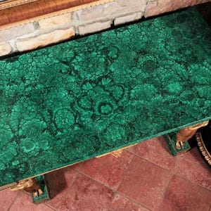 Puede incluir: Una mesa de centro rectangular con una tapa de patrón de malaquita verde vibrante. La mesa tiene detalles decorativos dorados en las patas y los bordes. Las dimensiones de la mesa no son visibles en la imagen.