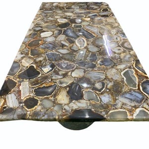 May include: A rectangular table top with a mosaic design. The surface is composed of irregularly shaped, polished agate stones in shades of gray, brown, and white. The stones are set in a resin base, creating a smooth, glossy finish.