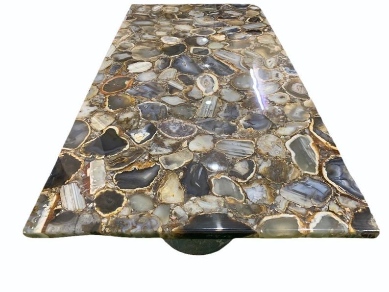 May include: A rectangular table top with a mosaic design. The surface is composed of irregularly shaped, polished agate stones in shades of gray, brown, and white. The stones are set in a resin base, creating a smooth, glossy finish.