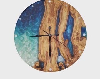 May include: Round wooden wall clock with a unique design. The clock face features a combination of natural wood grain and blue resin, creating a striking visual contrast. Black clock hands and hour markers complete the design.
