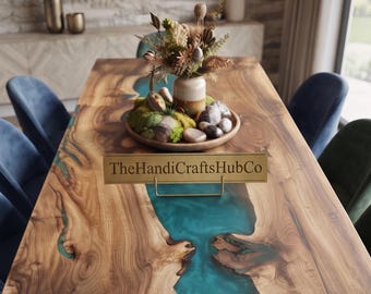 Può includere: Un tavolo da pranzo rettangolare con superficie in legno naturale e un intarsio centrale in resina traslucida color verde acqua. Un cartello in metallo dorato con la scritta TheHandiCraftsHubCo è posizionato vicino a una ciotola in legno con muschio e fiori secchi.