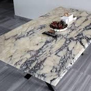 Op de afbeelding: Rechthoekige salontafel met een marmerlook oppervlak in wit, grijs en zwart. De tafel heeft zwarte poten en een decoratieve schaal met bessen en een afstandsbediening. De tafel is ongeveer 120 cm lang.