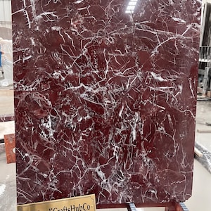 Pode incluir: Uma grande laje retangular de mármore vermelho profundo polido, apresentando uma rede intrincada de veios brancos brilhantes por toda a superfície. Uma pequena placa retangular de latão no canto inferior esquerdo exibe o texto TheHandiCraftsHubCo.