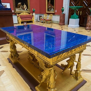 May include: A large, rectangular table with a vibrant blue top and ornate gold-colored supports. The table's surface reflects the surrounding room. The supports feature sculpted figures. The table is a striking piece of furniture.