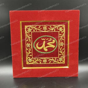Op de afbeelding: Een vierkant, rood fluwelen frame met een gouden rand omringt een bordeauxrood geborduurd paneel. Het paneel heeft gouden bloemmotieven en een ovaal met groene Arabische kalligrafie. Het frame is een decoratief stuk.