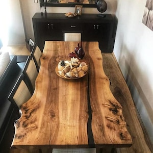 May include: A large, natural wood dining table with a live edge design. The table features a rich, brown color with unique wood grain patterns. A decorative bowl filled with seashells and starfish sits on the table. The table is surrounded by chairs with beige fabric seats.