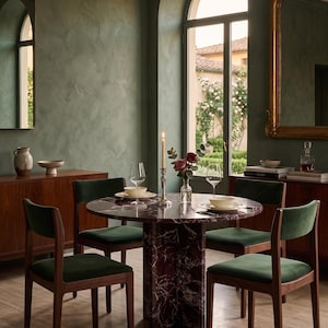 May include: A dining room set featuring a round marble table with a dark red and white pattern, surrounded by four wooden chairs with green velvet seats. The room has a green textured wall, a window, and a gold-framed mirror.
