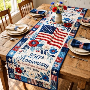 May include: A quilted table runner featuring a patriotic theme with red, white, and blue floral patterns, a central American flag design, and text reading 250th Anniversary The United States of America 1776-2026, accented by matching napkins.