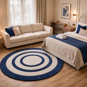 May include: A large circular area rug featuring a concentric pattern of alternating navy blue and cream rings. The rug is positioned on a light wood floor in a room containing a beige sofa, a bed with blue accents, and a side table with a lamp.