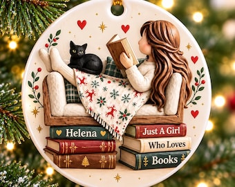 May include: A round ceramic ornament featuring a person with long brown hair reading a book on a sofa with a black cat. A blanket with reindeer patterns covers their legs. Below the sofa are books labeled Helena, Just A Girl, Who Loves, and Books.