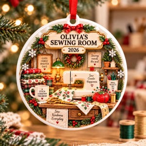 May include: A round white Christmas ornament featuring a sewing room scene with a sewing machine, fabric stacks, a mug reading A STITCH A BRIGHTER TOMORROW, a red pincushion, and text reading OLIVIA'S SEWING ROOM 2026, Sew Create Happiness, and Sew Kindness Everyday.