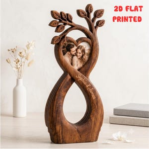 May include: A brown wooden infinity-shaped photo frame featuring a tree design at the top with leaves. The frame contains a heart-shaped photo of a couple and the engraved text "Sophia & James" on the side. The text "2D FLAT PRINTED" is visible.