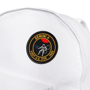 Puede incluir: Mochila blanca con un parche circular. El parche tiene el texto "Gemini 4" y "First U.S. EVA 1965". Muestra un astronauta con traje espacial, un planeta rojo y una nave espacial. La mochila está hecha de un material duradero.