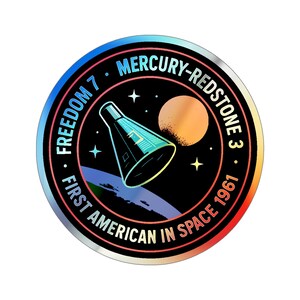 May include: A circular sticker with a holographic finish. The design features a space capsule, a planet, stars, and the text "FREEDOM 7", "MERCURY-REDSTONE 3", "FIRST AMERICAN IN SPACE 1961". The sticker has a black background with a blue, red, and orange border.