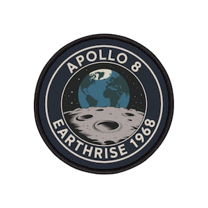 Może przedstawiać: Okrągła naszywka upamiętniająca misję Apollo 8. Projekt przedstawia Ziemię wschodzącą nad powierzchnią Księżyca, z napisem "APOLLO 8 EARTHRISE 1968" w okręgu. Naszywka ma granatowe tło z białymi i jasnoniebieskimi akcentami.