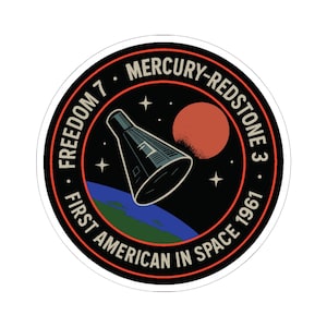 May include: A round sticker featuring a retro space-themed design. The sticker has a black background with a rocket ship, a red planet, stars, and the Earth. Text includes "Freedom 7", "Mercury-Redstone 3", and "First American in Space 1961".