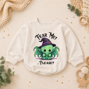 May include: A cream-colored long-sleeve sweatshirt featuring a graphic of a small green creature with wings and a purple witch hat. The text above the creature reads "FEAR ME!" and the text below reads "...PLEASE?" in a black, stylized font.