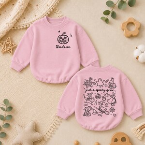 May include: Two light pink long-sleeve sweatshirts. The top one features a small black pumpkin graphic and the name Hudson. The bottom one displays a large black graphic print of various geese in costumes with the text just a spooky goose.