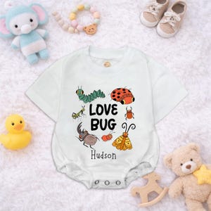 Op de afbeelding: Witte babyromper met een kleurrijk insectenontwerp en de tekst "LOVE BUG" met de naam "Hudson" eronder. Het ontwerp bevat een lieveheersbeestje, rups en andere insecten. Babyspeelgoed omringt de romper.