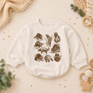 May include: A cream-colored long-sleeve sweatshirt featuring a brown graphic print of multiple raccoons in various poses. The garment has a crew neckline, ribbed cuffs, and a gathered hemline, showcasing detailed illustrations of the animals.