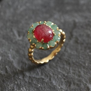 Op de afbeelding: Een gouden ring met een centrale ovale rode edelsteen, omringd door groene edelstenen. De band van de ring heeft een gestructureerd, kralenontwerp. De ring ligt op een donkergrijze, getextureerde ondergrond.
