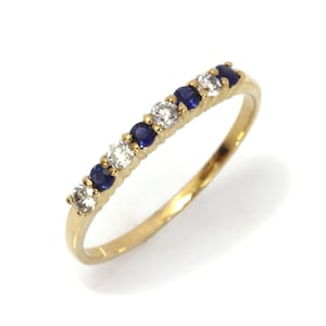 May include: A gold ring featuring alternating round, brilliant-cut white and blue gemstones. The ring's band is a polished gold color, and the gemstones are set in a row across the top. The ring is set against a white background.