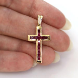 Puede incluir: Un colgante de oro con forma de cruz, adornado con piedras preciosas rojas de corte cuadrado. La cruz tiene un diseño sencillo y elegante con un lazo en la parte superior para una cadena. Las piedras preciosas están engastadas en un engaste de canal, creando una línea continua de color.