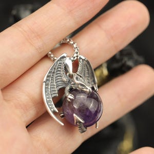 May include: A silver-colored dragon pendant necklace. The dragon has detailed wings and is clutching a large, round, purple amethyst stone. The necklace chain is also silver. The pendant is held in a hand.