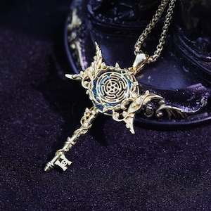 May include: Gold-coloured pendant necklace with a key design. The pendant has a central circular design with a blue background, surrounded by ornate floral and leaf details. The key's handle is intricately designed, and the chain is a delicate link style.