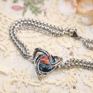 May include: A silver-toned bracelet featuring a double box chain and a central pendant. The pendant consists of an intricate metal knot design surrounding a circular iridescent blue and purple stone with a small, triangular orange gem.