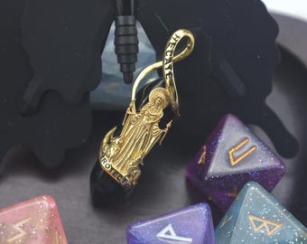May include: A gold-colored pendant with the word "HECATE" and a figure of a woman. The pendant is attached to a black stone. Several polyhedral dice in shades of purple, blue, and pink are in the background.