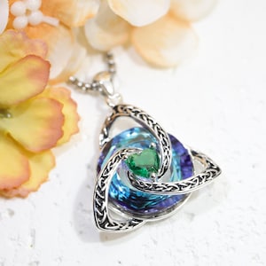 Può includere: Un ciondolo color argento con una cornice triangolare a nodo celtico che circonda un cristallo circolare iridescente blu e viola. Un piccolo cristallo verde a forma di cuore è incastonato al centro della pietra vorticosa multicolore.