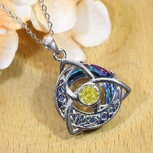 Pode incluir: Um colar com pingente em tom prateado apresentando um design de nó triquetra circular com trabalho em metal intrincado. O centro contém uma pedra preciosa amarela redonda e facetada, enquanto o fundo do nó exibe um acabamento arco-íris iridescente.