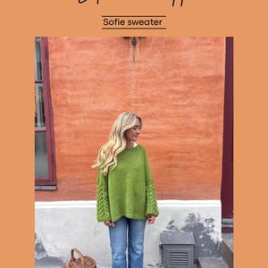 May include: A woman wearing a green knit sweater and blue jeans stands in front of an orange wall. The sweater has cable knit detailing on the sleeves. The image includes the text "Strikkerhopp" and "Sofie sweater". A woven basket sits on the ground.