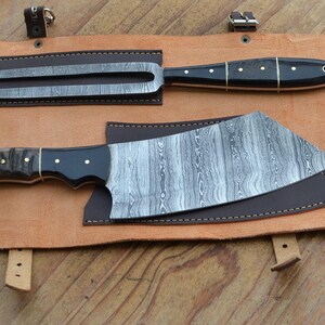 May include: A set of two Damascus steel knives with black handles and brass accents, resting on a tan leather sheath. The set includes a cleaver and a carving fork, ideal for culinary use.