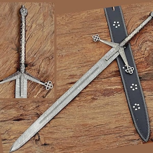 Könnte beinhalten: Ein detailliertes Bild eines langen, verzierten Schwertes mit silbernem Griff und Parierstange. Die Klinge hat ein gemustertes Design. Das Schwert liegt auf einer Holzoberfläche neben einer schwarzen Lederscheide mit weißen Tupfen. Der Griff des Schwertes hat ein kronenartiges Design.