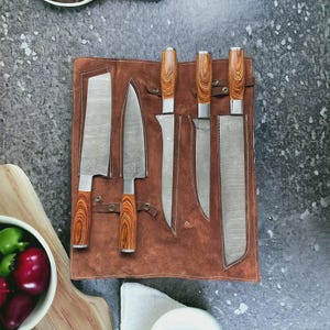 Puede incluir: Un juego de cinco cuchillos de cocina de acero inoxidable con mangos de madera, exhibidos en un estuche de cuero marrón. Las hojas tienen un diseño estampado. Los mangos son de color marrón claro con un patrón de vetas de madera.