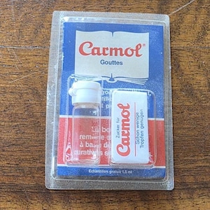 Può includere: Un piccolo flacone di vetro trasparente con un tappo bianco e una piccola confezione rettangolare, entrambi etichettati "Carmol". La confezione è blu e bianca, con il marchio in rosso. Il flacone contiene un liquido rosso. Il testo "Gouttes" è visibile.