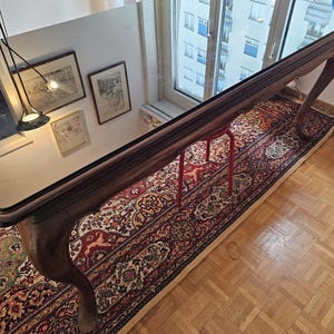 Puede incluir: Una mesa larga de madera oscura con tapa de cristal, con patas curvas y ornamentadas. La mesa se asienta sobre una alfombra estampada con tonos rojos y crema. Una banqueta roja está colocada debajo. La habitación tiene pisos de madera y una ventana.