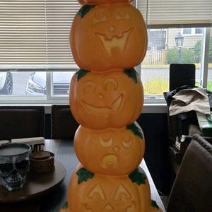May include: A vertical stack of five orange plastic jack-o'-lanterns with carved facial expressions and green leaf accents. The pumpkins are arranged in a column, with the largest pumpkin at the base and smaller ones stacked on top, creating a tall, singular decorative Halloween figure.