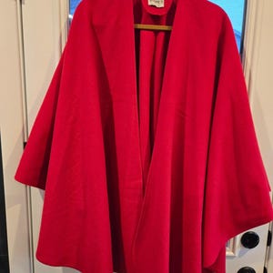 May include: A vibrant red cape hanging on a wooden hanger. The cape is made of a soft material and has a wide, open front. The label reads "JAMES".