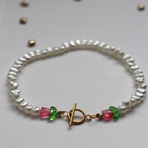 Puede incluir: Un delicado collar de perlas con un cierre dorado y detalles florales decorativos rosas y verdes. El collar está compuesto por pequeñas perlas blancas y presenta un cierre de palanca. Los detalles florales son rosas y verdes.