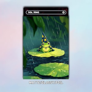 May include: A rectangular trading card titled SOL RING featuring an illustration of a green frog wearing a blue wizard hat with yellow stars, sitting on a lily pad in the rain. The text MATTE/GLOSSY/FOIL is printed at the bottom of the card.
