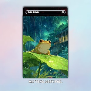 Puede incluir: Carta con una rana verde sobre una hoja bajo la lluvia. El texto "SOL RING" está en la parte superior y "MATTE/GLOSSY/FOIL" en la inferior. La rana tiene ojos naranjas y está sobre una hoja verde. El fondo muestra un edificio.