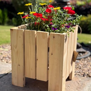 May include: A wooden planter filled with red, yellow, and purple flowers. The planter is made from light-coloured wooden planks, with a natural, rustic look. The flowers are in full bloom, creating a vibrant display of colour. The planter is outdoors.