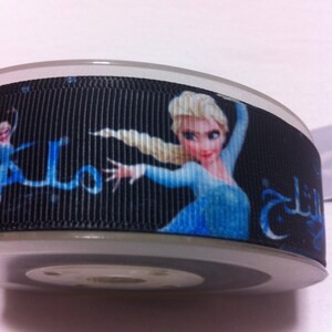 May include: Black ribbon featuring a cartoon image of a character in a blue dress with blonde hair. The ribbon has text in a foreign language. The ribbon is wrapped around a spool.