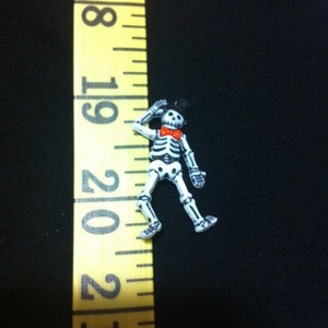 May include: A white skeleton figurine wearing a black top hat and a red bow tie. The figurine is about 1 inch tall and is standing on a black surface.